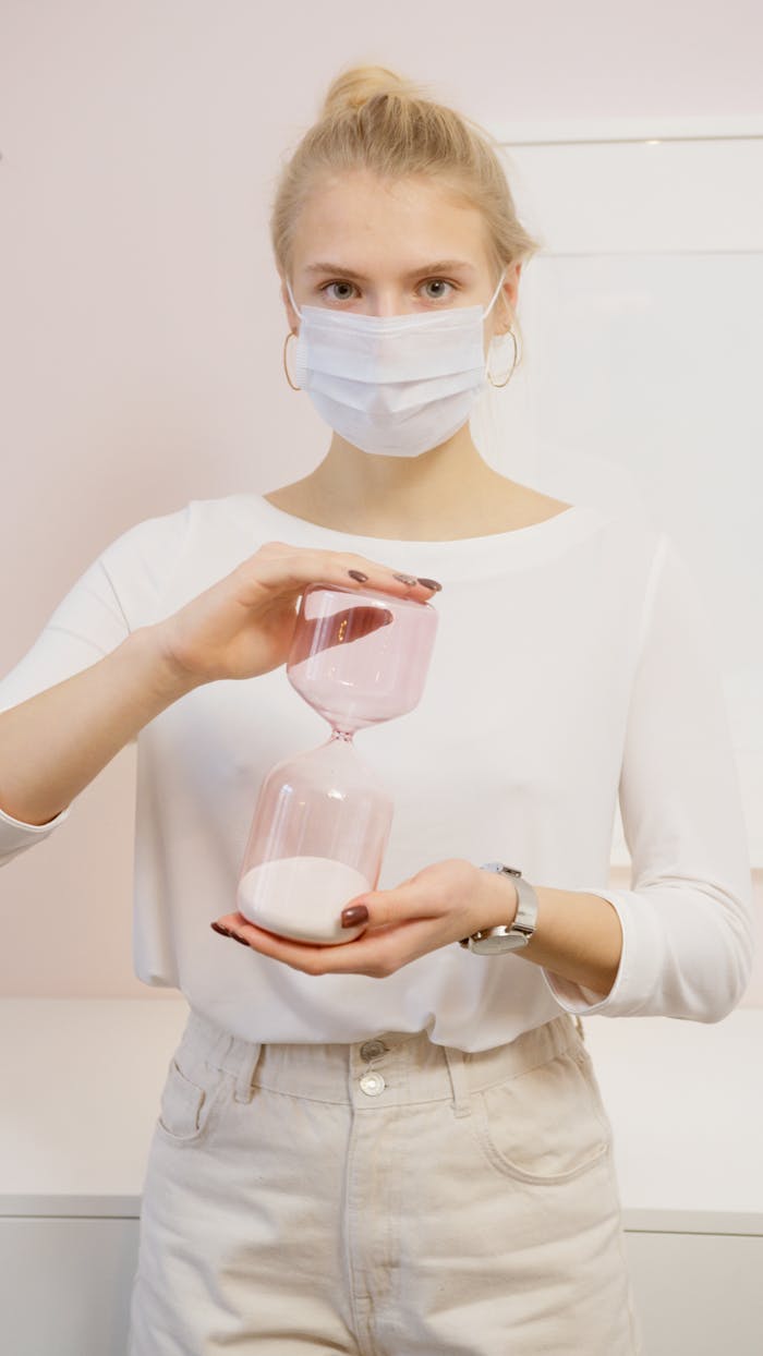 A woman in white holding an hourglass with a protective facemask, symbolizing time and health.